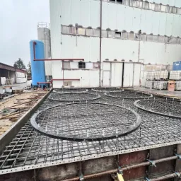 Zbrojenie fundamentu pod konstrukcję przemysłową z widocznymi okrągłymi elementami wzmacniającymi i szalunkiem, na tle budynku przemysłowego i zbiorników.