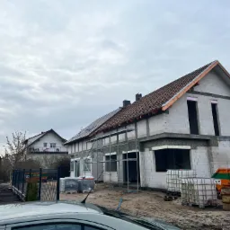 Budowa domu murowanego z białych bloczk&oacute;w, widoczne rusztowania, okna bez szyb, dach pokryty dach&oacute;wką, kontener EKO-TOM na gruz.