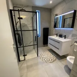 Nowoczesna łazienka z czarną kabiną prysznicową, białymi płytkami marmuropodobnymi, podświetlanym lustrem i okrągłym dywanikiem z geometrycznym wzorem.