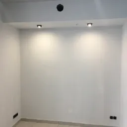 Pomalowane na biało wnętrze pokoju z szarą podłogą z płytek, widoczne gniazdka elektryczne, trzy małe lampy w suficie podwieszanym i otwór na oświetlenie.