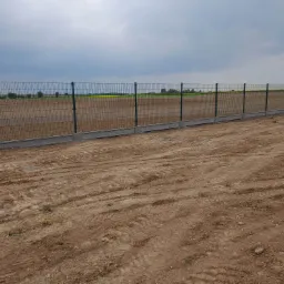 Długi, stalowy płot z paneli na betonowej podmurówce, oddzielający pole uprawne od piaszczystego terenu z widocznymi śladami opon, pod pochmurnym niebem.