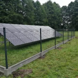 Panel słoneczny otoczony ciemnozielonym ogrodzeniem z siatki na betonowej podmurówce, na tle zielonej trawy i drzew