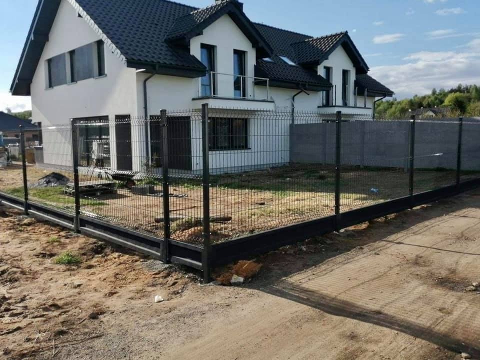 Nowoczesne ogrodzenie panelowe w kolorze grafitowym, zamontowane wokół domu z białymi ścianami i ciemnym dachem, widok z zewnątrz, teren jeszcze niezagospodarowany.