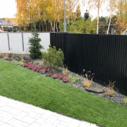 Nowoczesny ogród z równo przystrzyżonym trawnikiem, rabatą z wrzosami i ozdobnymi trawami, wykończoną szarym kamieniem i czarnym, pionowym ogrodzeniem.