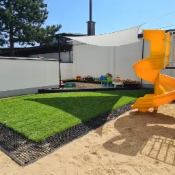 Widok na plac zabaw z nowo położoną trawą w rolkach, oddzieloną od piasku czarną obwódką, z białym płotem i markizą w tle.