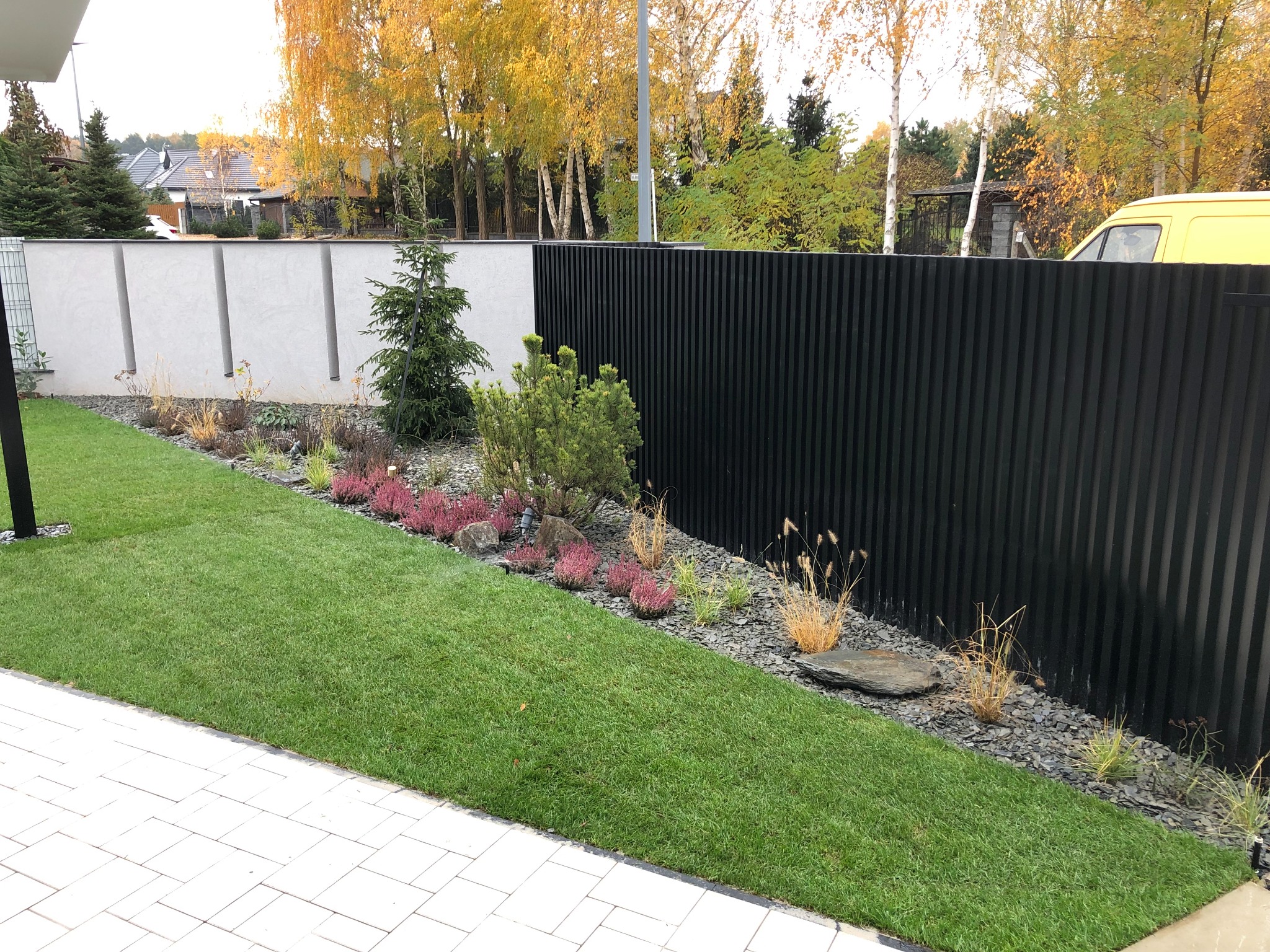 Nowoczesny ogród z równo przystrzyżonym trawnikiem, rabatą z wrzosami i ozdobnymi trawami, wykończoną szarym kamieniem i czarnym, pionowym ogrodzeniem.