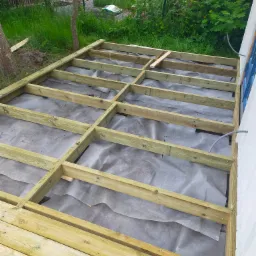 Konstrukcja drewnianego tarasu w trakcie budowy, legary ułożone na szarej geowłókninie, widoczne elementy podwaliny przy ścianie budynku i fragment ogrodu w tle.