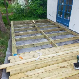 Budowa drewnianego tarasu: widoczna konstrukcja ramy z legarów na szarej membranie, przygotowane deski tarasowe, nóż tapicerski i kątownik w tle biały budynek z niebieskimi drzwiami.