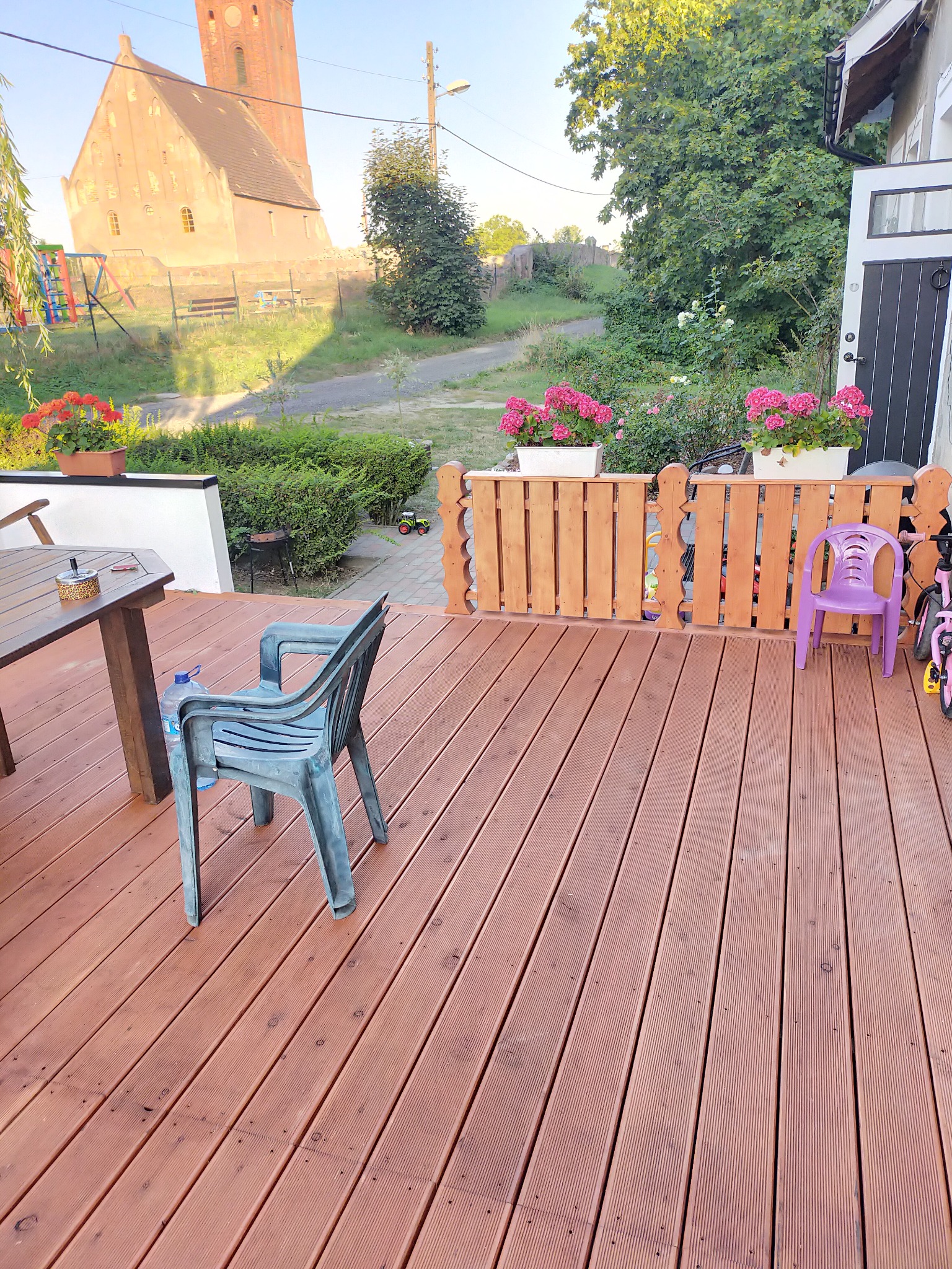Drewniany taras z widokiem na kościół, plastikowe krzesła, stół z popielniczką, kwiaty w donicach na ażurowym ogrodzeniu, zabawki dziecięce w tle.