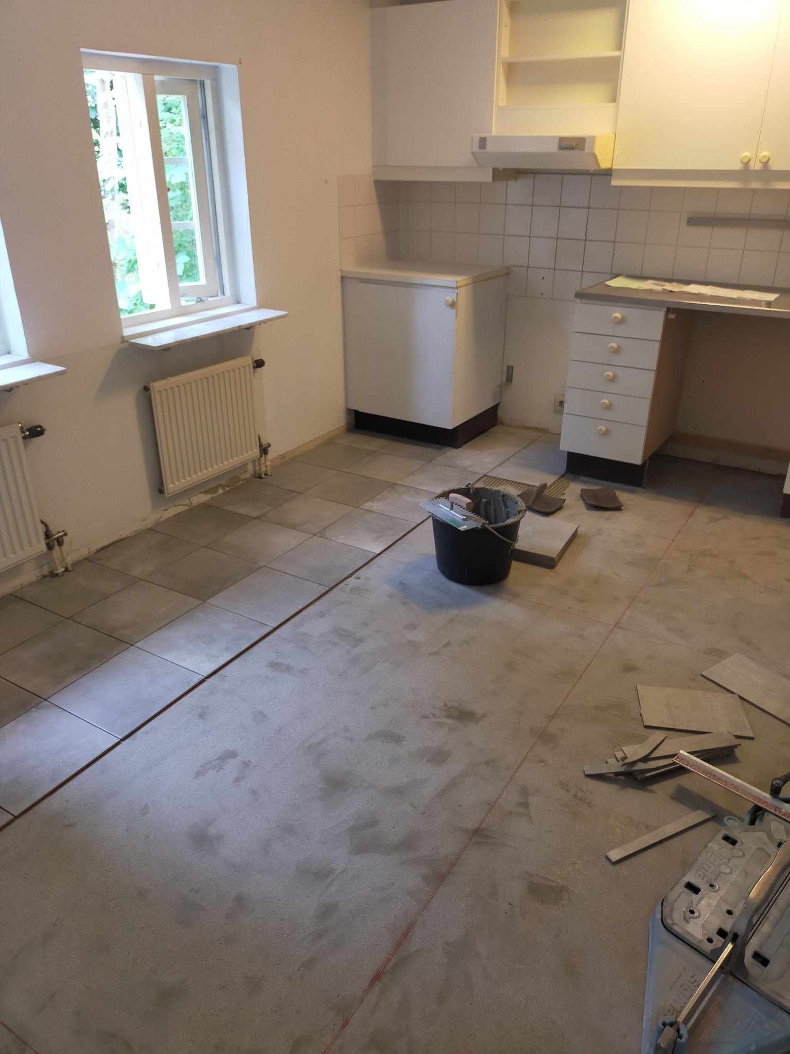Układanie szarych płytek podłogowych w kuchni, widoczny kubeł z zaprawą, przycięte płytki, oraz maszynka do cięcia płytek.