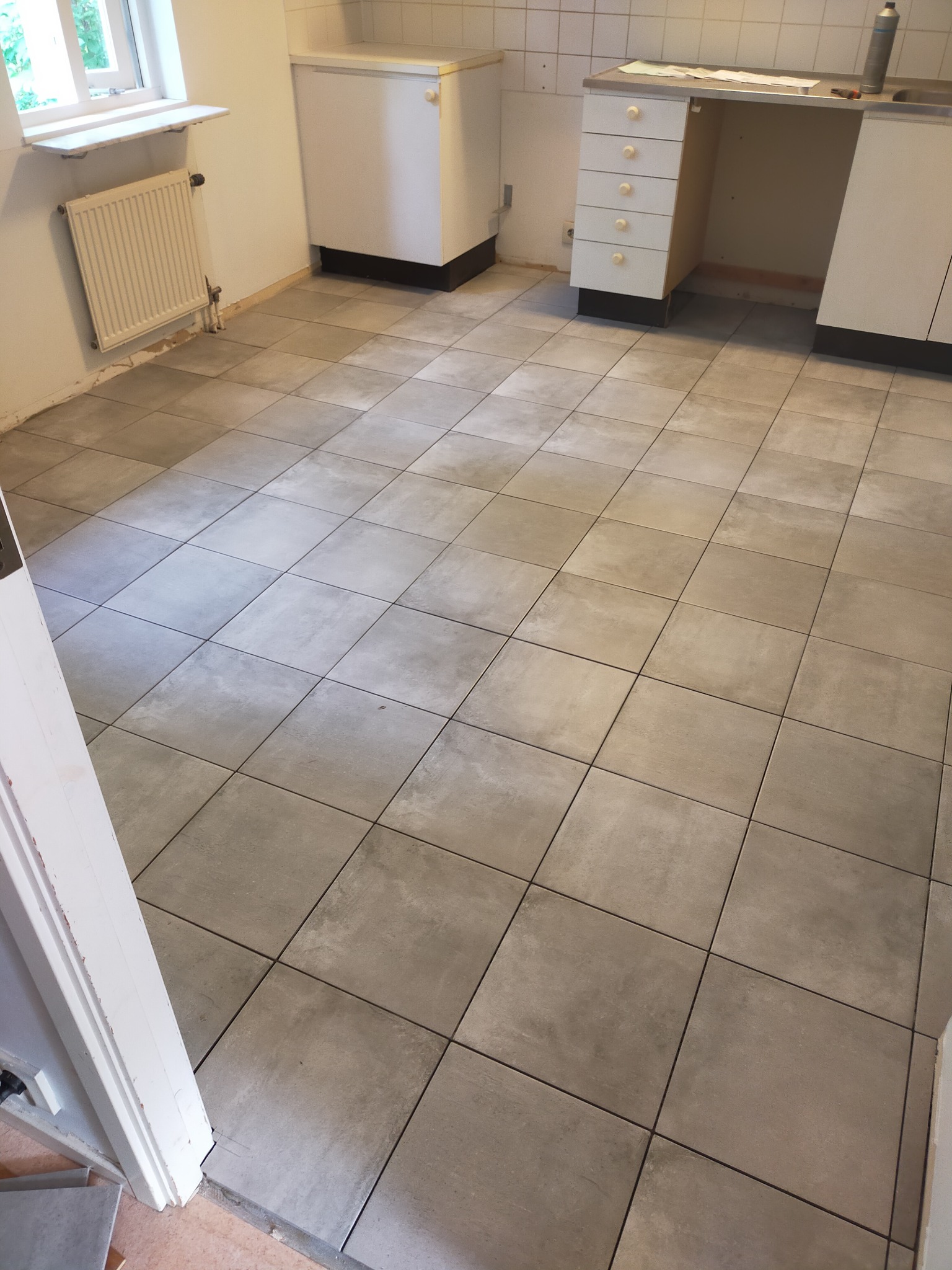 Ułożona szara, kwadratowa terakota podłogowa w kuchni, widoczne białe szafki kuchenne, grzejnik i fragment ościeżnicy drzwiowej.