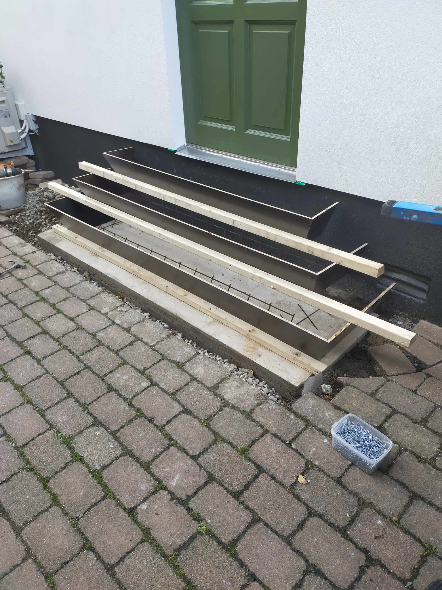 Przygotowanie do wylewki betonowej schodów wejściowych: szalunek z desek, zbrojenie i pojemnik ze śrubami na kostce brukowej obok budynku z zielonymi drzwiami.