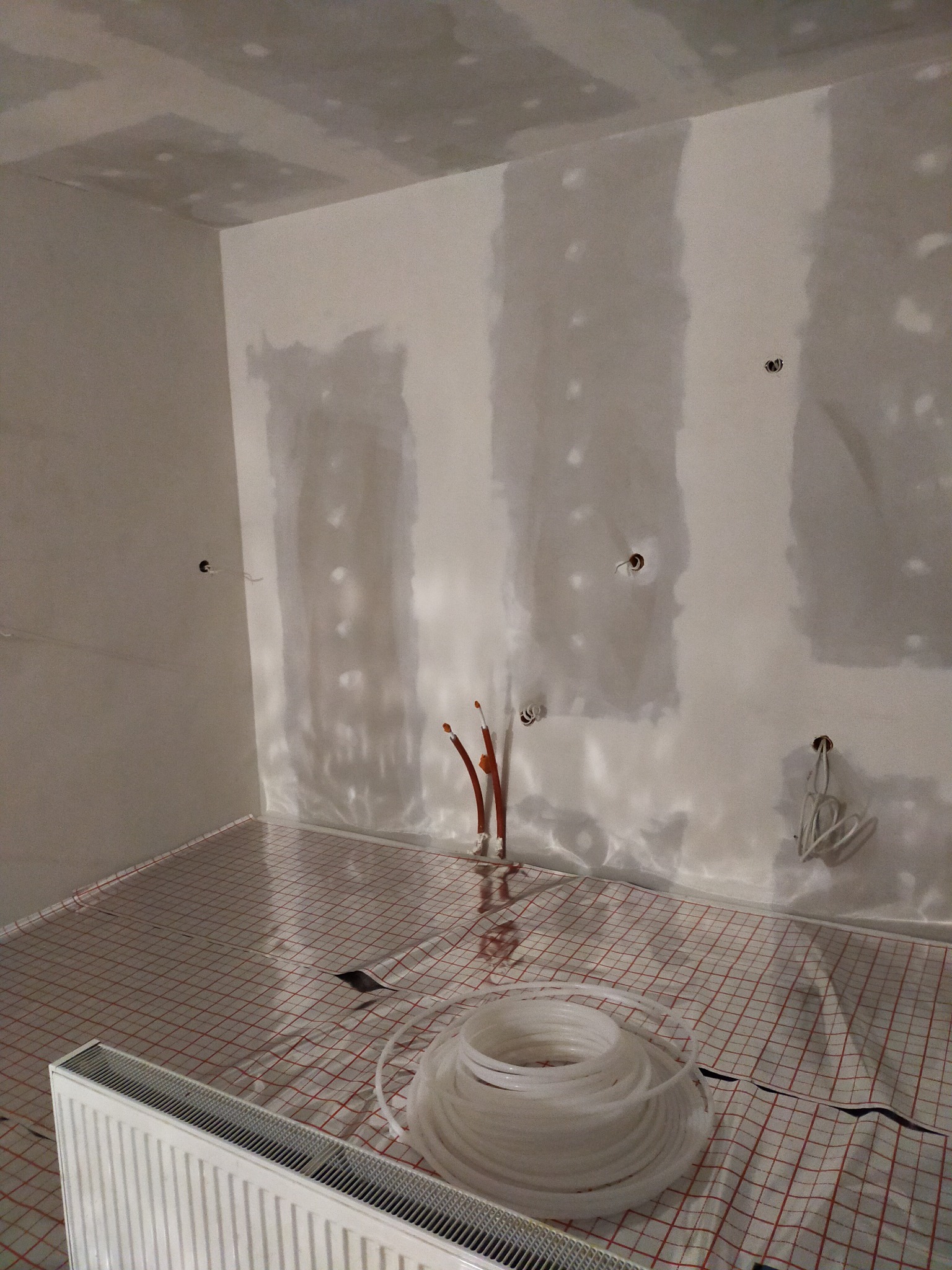 Wnętrze w trakcie prac wykończeniowych: ściany z płyt kartonowo-gipsowych wymagające szpachlowania, instalacja ogrzewania podłogowego z rurami na podłodze, widoczny grzejnik i wystające rury...