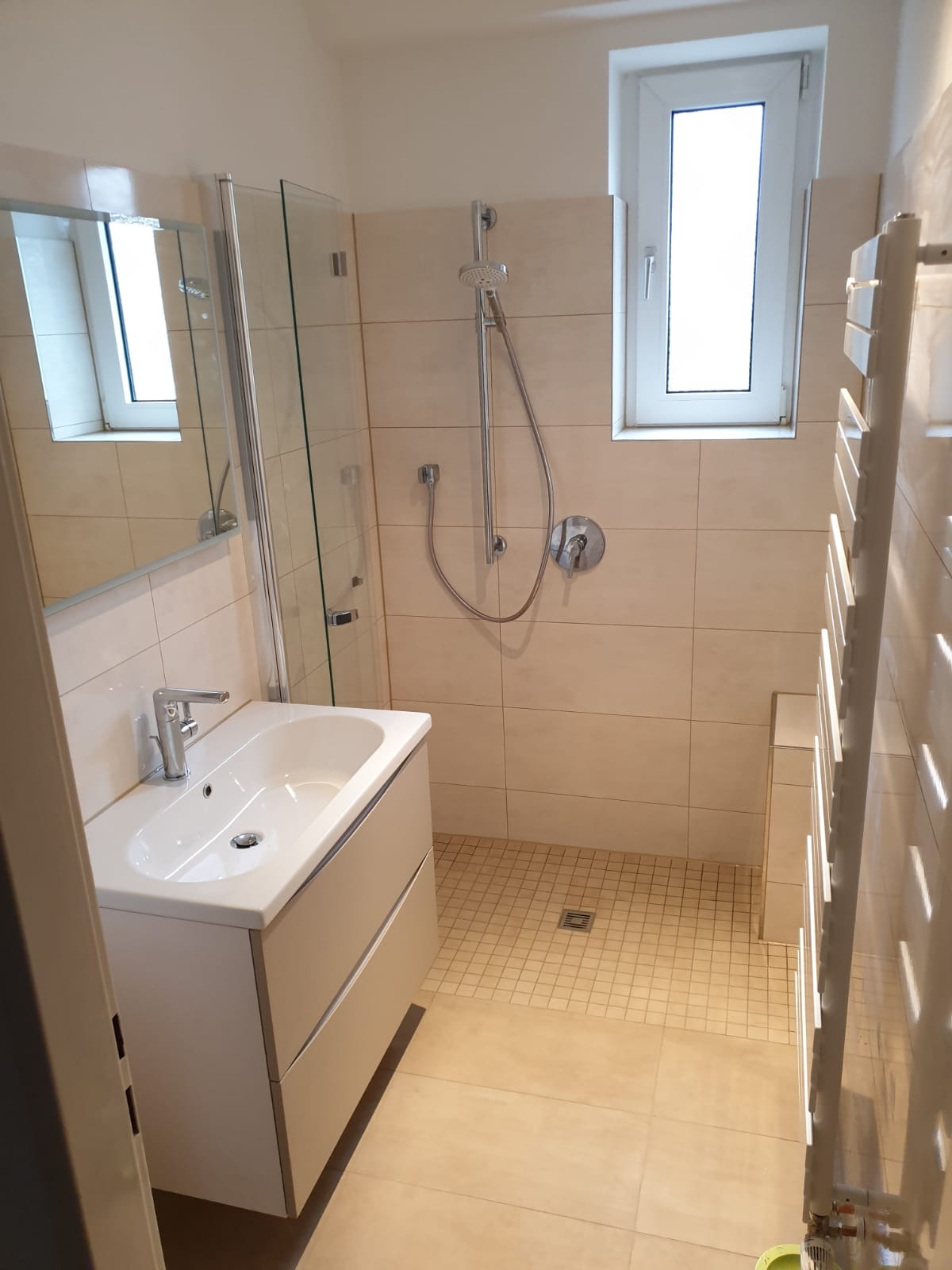 Jasna łazienka po remoncie z beżowymi płytkami, kabiną prysznicową, umywalką z szafką i grzejnikiem drabinkowym.