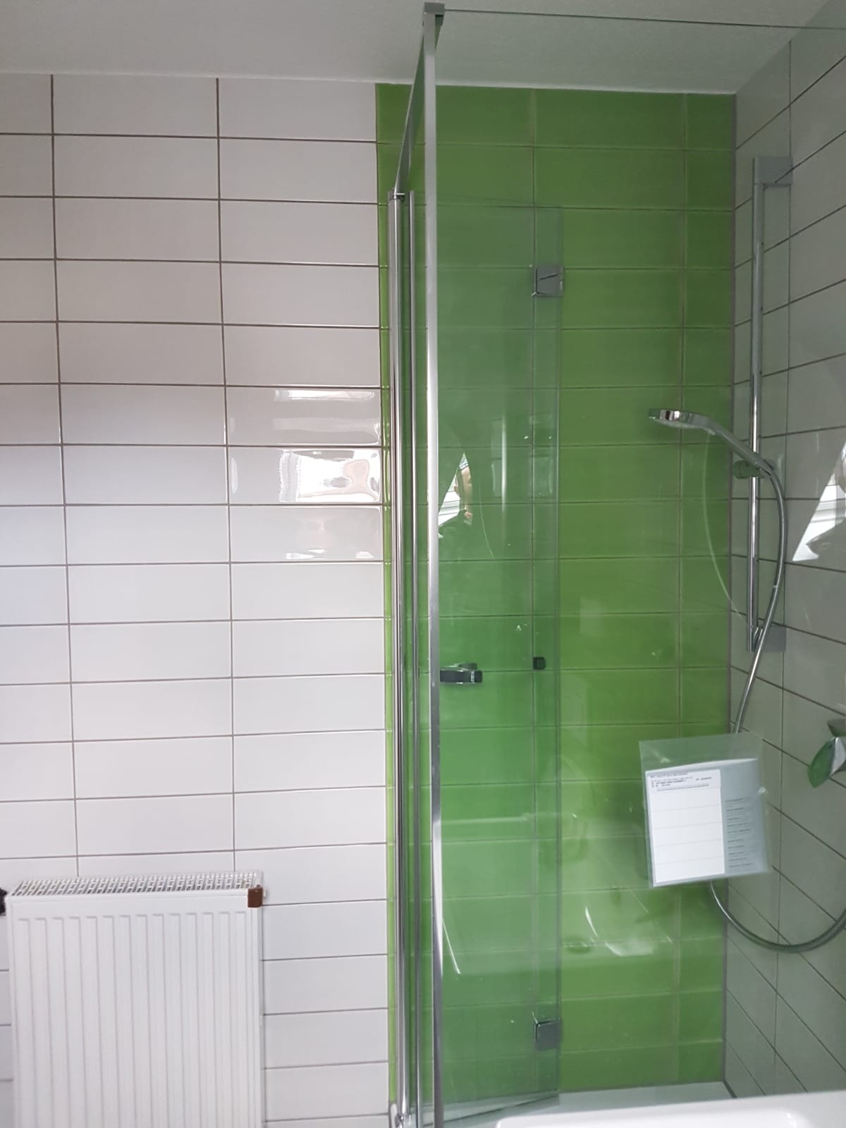 Kabina prysznicowa z zielonymi i białymi płytkami, widoczny grzejnik i dokumentacja techniczna wewnątrz kabiny.