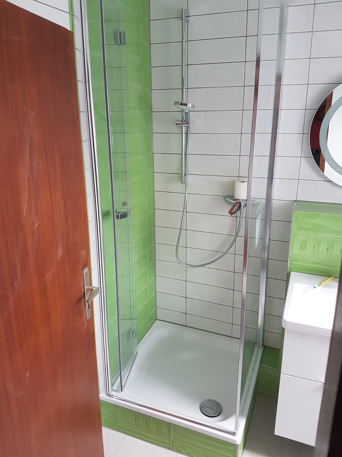 Nowoczesna kabina prysznicowa z transparentnymi drzwiami, zielone i białe płytki na ścianach, biała umywalka z żółtą szczoteczką do zębów, okrągłe lustro z podświetleniem, brązowe drzwi wejściowe.