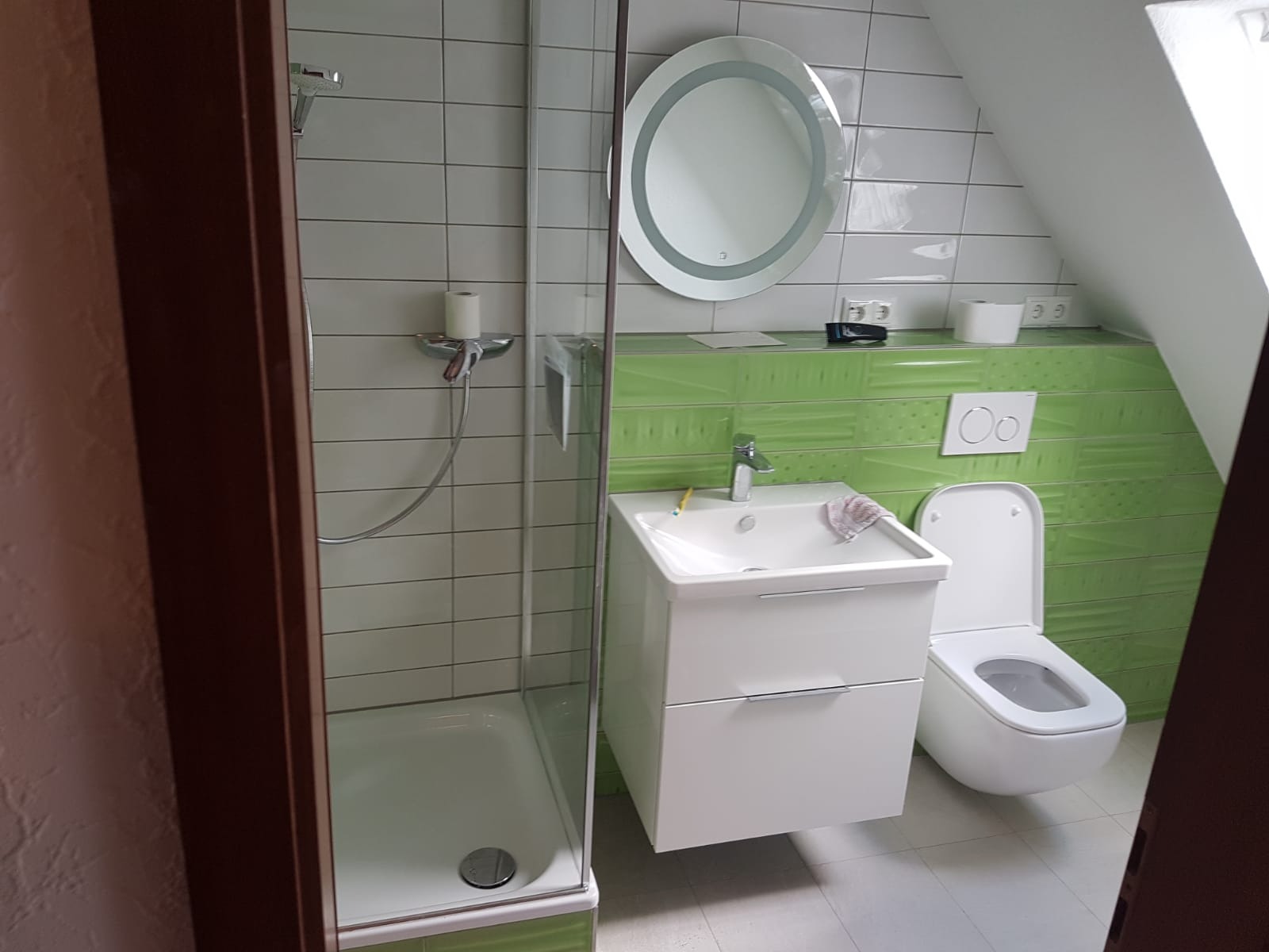 Wyremontowana łazienka na poddaszu z kabiną prysznicową, białą umywalką z szafką, toaletą i okrągłym lustrem z podświetleniem, wykończona zielonymi i białymi płytkami.