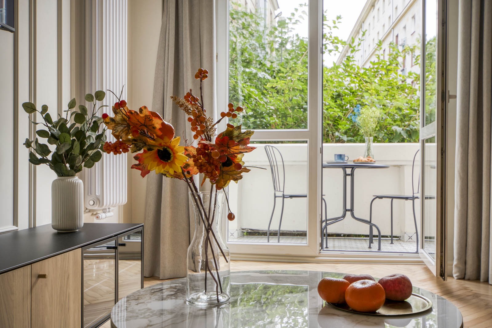 Jasne wnętrze mieszkania z otwartym balkonem, na marmurowym stoliku owoce, w tle widok na zielone drzewa i budynki.