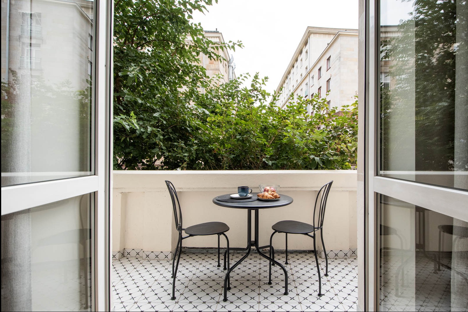 Klimatyczny balkon z czarno-białą mozaiką podłogową, metalowym stolikiem z filiżanką i rogalikami oraz widokiem na zieleń drzew między budynkami.