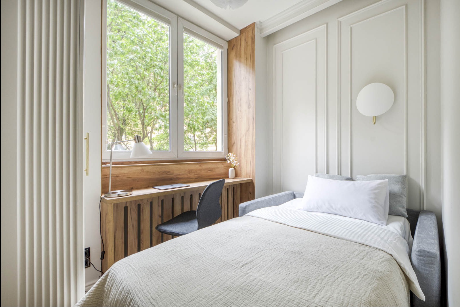 Jasne wnętrze pokoju z łóżkiem, biurkiem z drewnianym blatem pod oknem i szarą tapicerowaną ścianą z dekoracyjnymi listwami oraz okrągłym kinkietem