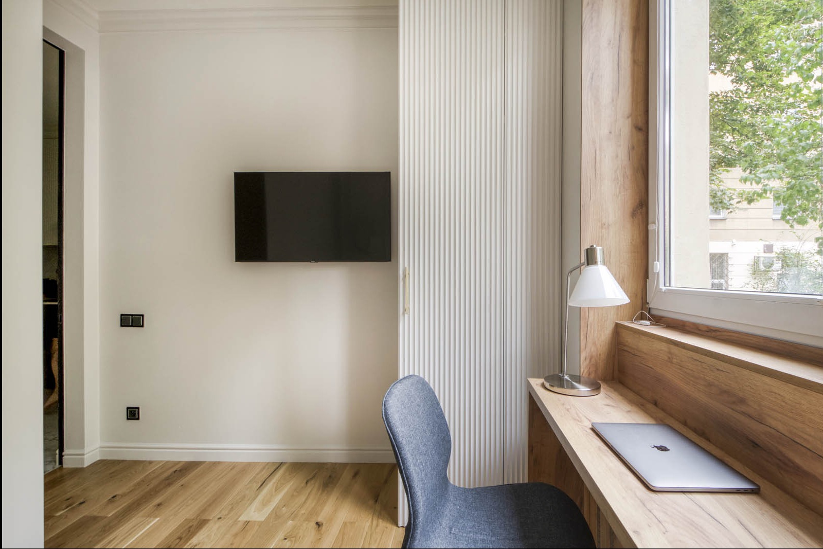 Nowoczesny pokój z drewnianą podłogą, biurkiem z laptopem przy oknie, minimalistycznym krzesłem i telewizorem na ścianie obok białej szafy z ryflowanymi drzwiami.