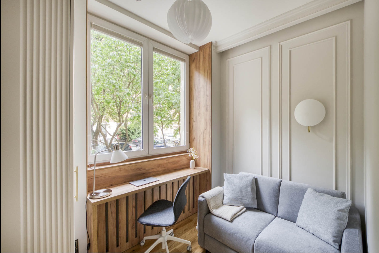 Jasne wnętrze pokoju z drewnianym biurkiem wbudowanym w okno, szarą sofą i dekoracyjnymi panelami ściennymi w stylu skandynawskim.