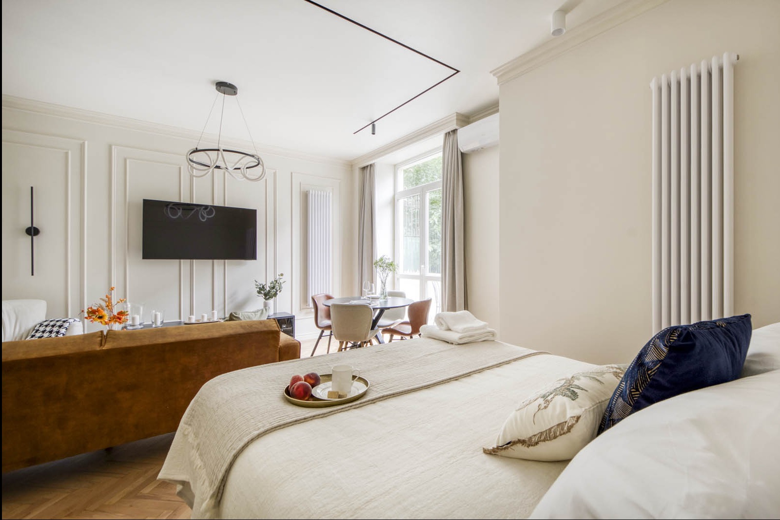 Jasne wnętrze mieszkania w stylu eklektycznym: widok na salon z łóżka, dekoracyjne panele ścienne, telewizor, okrągły stół jadalny i designerska lampa wisząca.