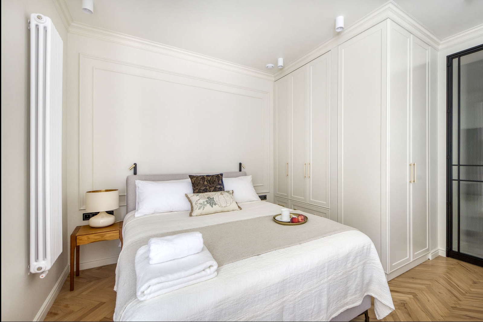 Jasna sypialnia z łóżkiem, drewnianą podłogą w jodełkę i białymi szafami w zabudowie, z widocznym dekoracyjnym grzejnikiem.