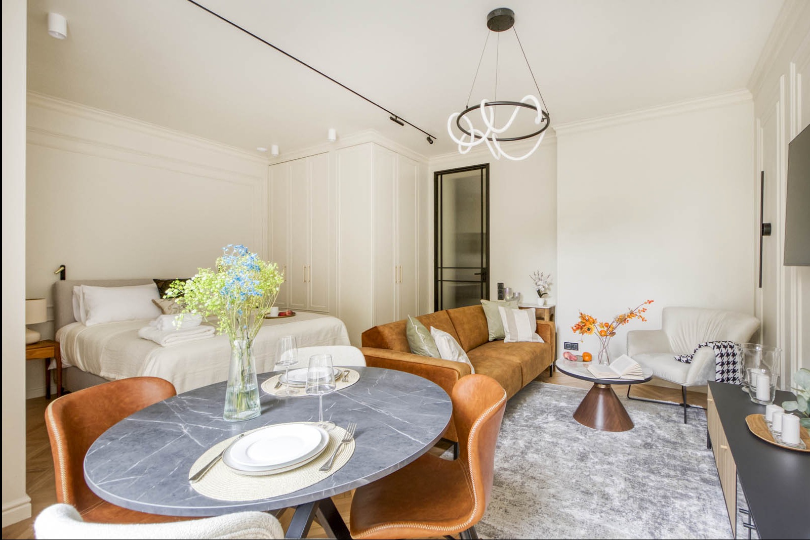 Jasne, nowocześnie urządzone studio: okrągły stół z marmurowym blatem, skórzane krzesła, brązowa sofa, białe szafy w zabudowie, dekoracyjne oświetlenie sufitowe, dywan w odcieniach szarości, kwiaty...