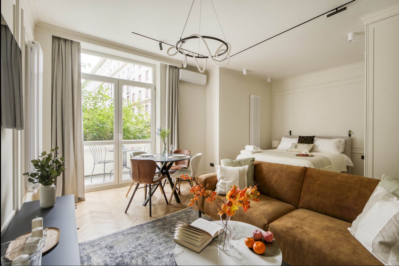 Jasne studio z parkietem w jodełkę, brązową sofą, marmurowym stolikiem kawowym z książką i owocami, okrągłym stołem jadalnym z krzesłami, podwójnym łóżkiem w tle i wyjściem na balkon.