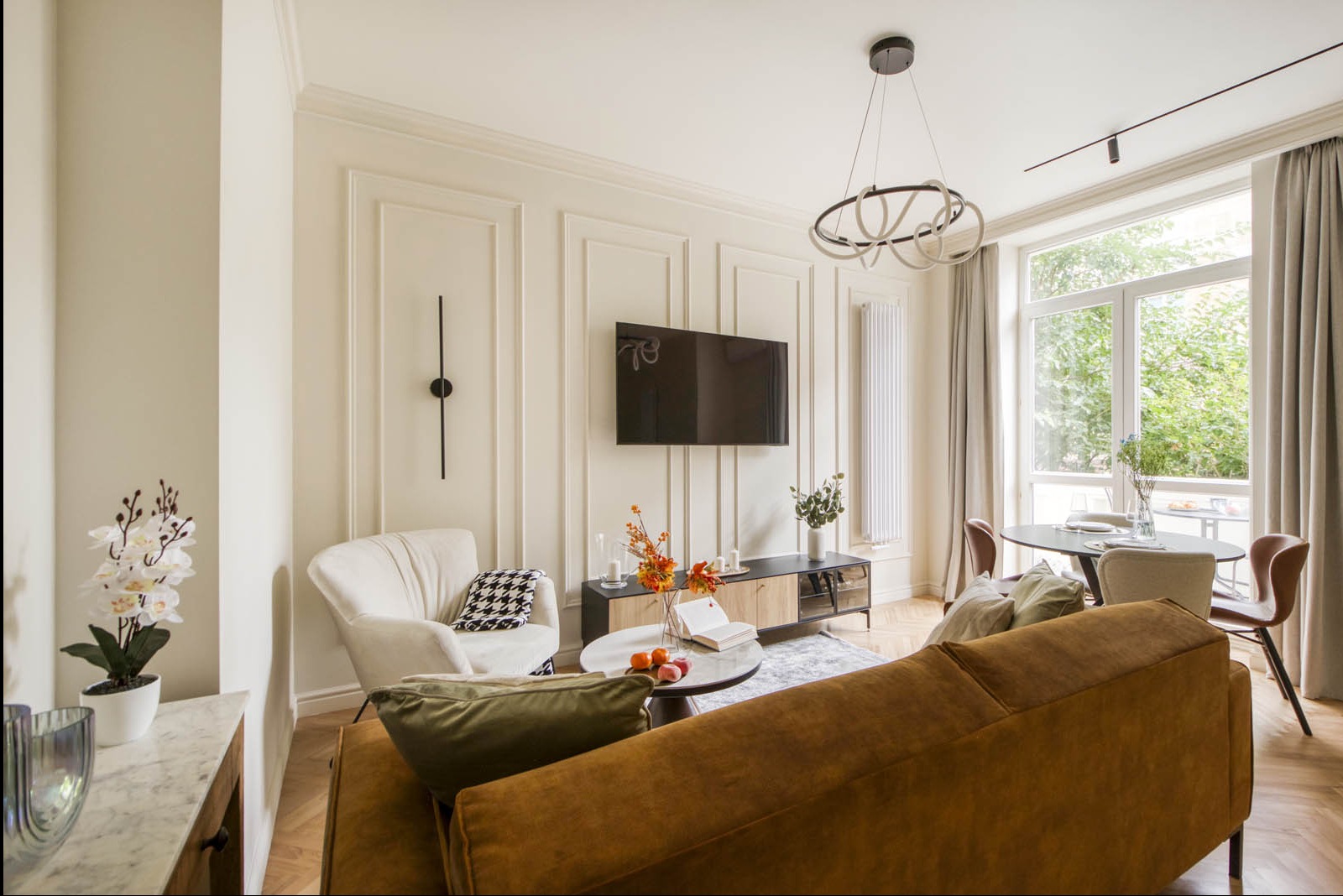 Elegancki salon w Warszawie z kremowymi ścianami zdobionymi sztukaterią, wiszącym telewizorem, nowoczesnym oświetleniem i parkietem w jodełkę. Widoczny fragment sofy w kolorze musztardowym, fotel...