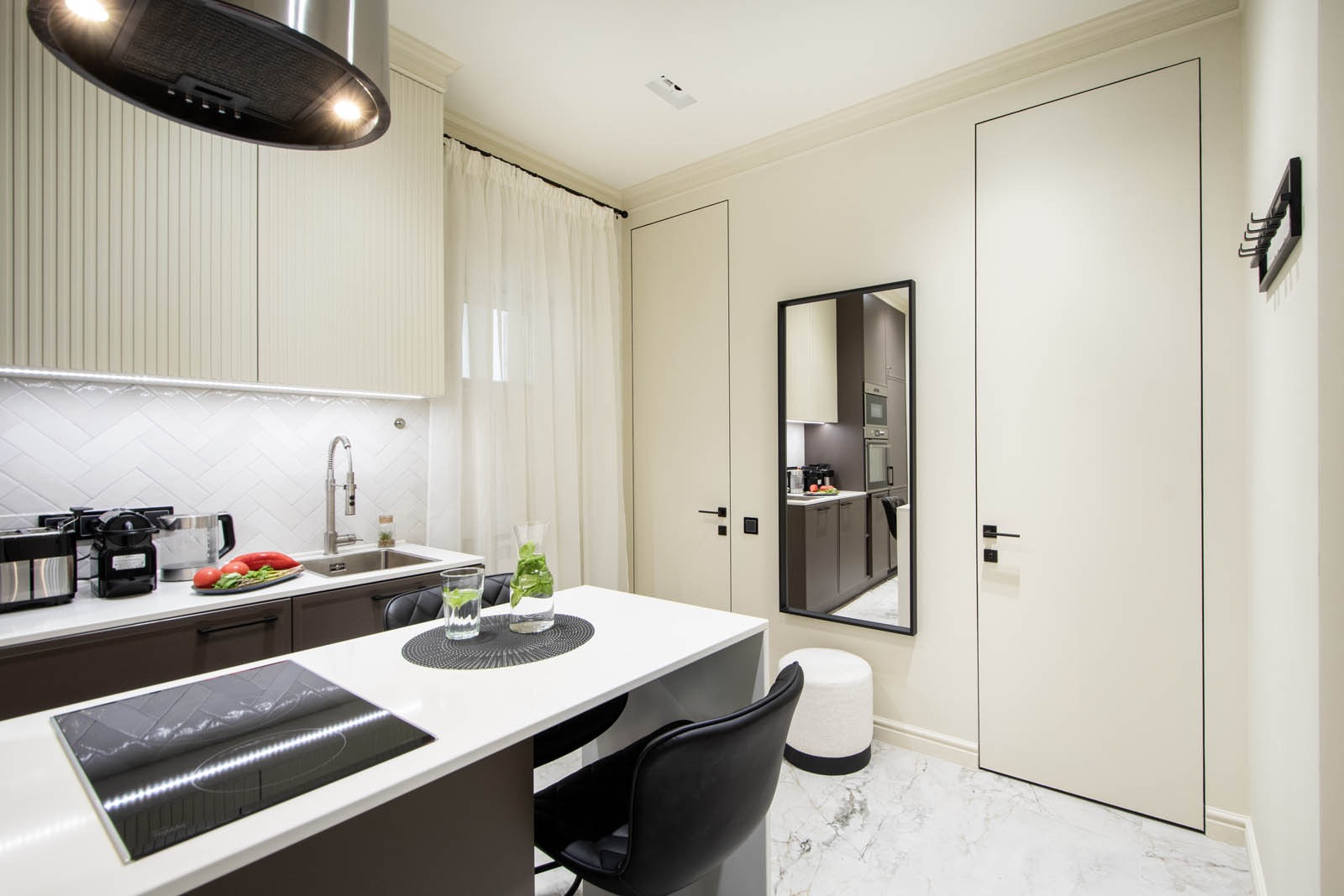 Nowoczesna kuchnia z marmurową podłogą, białymi szafkami z ryflowanymi frontami i ciemnobrązowymi dolnymi szafkami, wyspą kuchenną z indukcyjną płytą, lustrem odbijającym dalszą część kuchni...