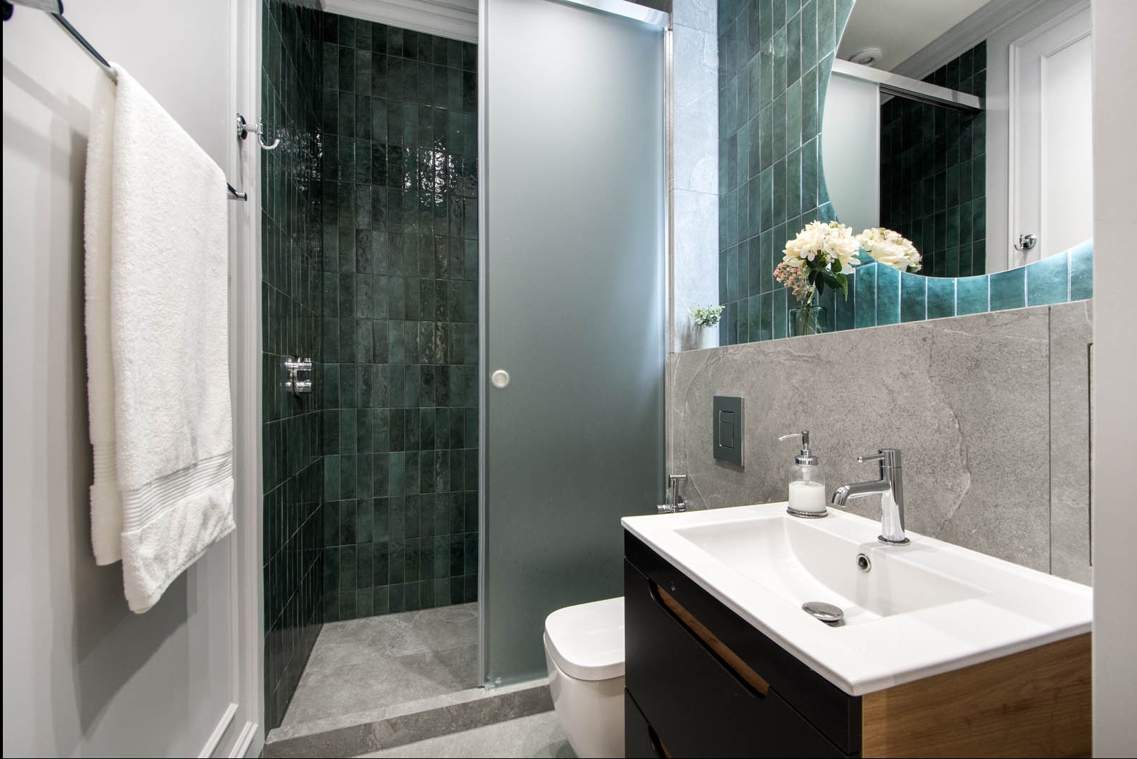 Nowoczesna łazienka z kabiną prysznicową walk-in wyłożoną ciemnozielonymi płytkami, umywalką nablatową z czarną szafką i szarą ścianą, oraz białym ręcznikiem wiszącym obok.