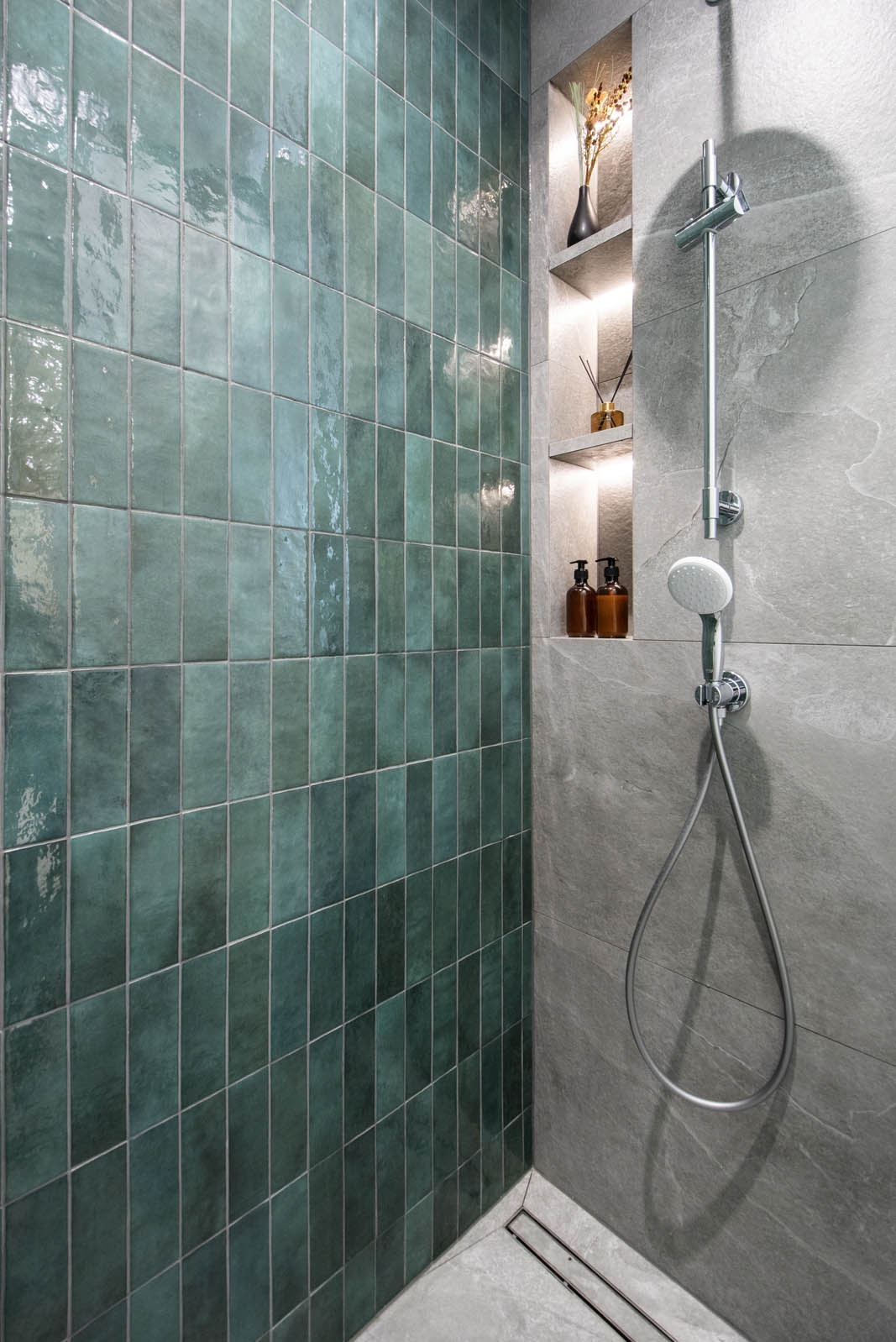 Kabina prysznicowa z zielonymi, pionowo ułożonymi płytkami o błyszczącej powierzchni, szare płytki imitujące kamień, wnęka z podświetleniem, dekoracje i dozowniki na kosmetyki.