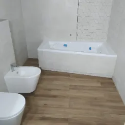 Nowoczesna łazienka z wanną obudowaną płytkami z fakturą, białą armaturą (bidet i toaleta) oraz podłogą imitującą drewno.