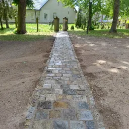 Ułożona z kamiennej kostki ścieżka prowadzi przez trawnik do bramki w ogrodzeniu przed domem. Widoczne drzewa i latarnia. Kostka brukowa w odcieniach szarości i beżu.