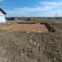 Wykonany wykop pod fundamenty z drewnianym szalunkiem, widok z góry na brązową ziemię i błękitne niebo. W tle budynek w stanie surowym i sterta ziemi.
