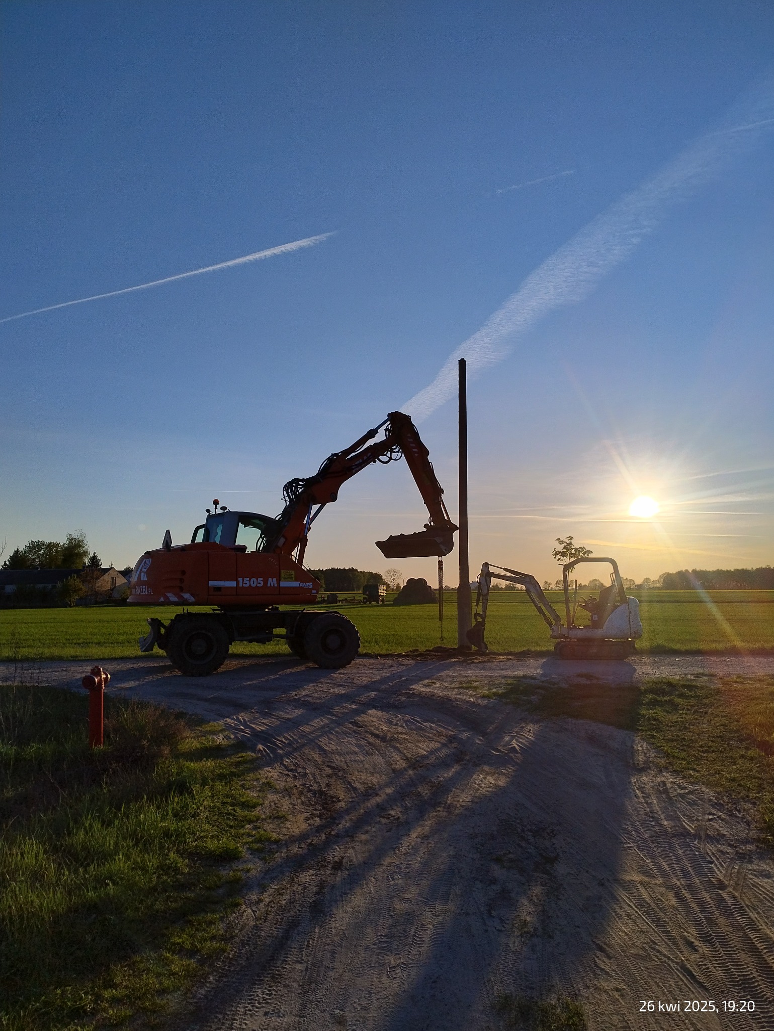 Dwie koparki (pomarańczowa i biała) pracują na polu przy słupie w promieniach zachodzącego słońca, widoczne ślady opon na piaszczystej drodze.
