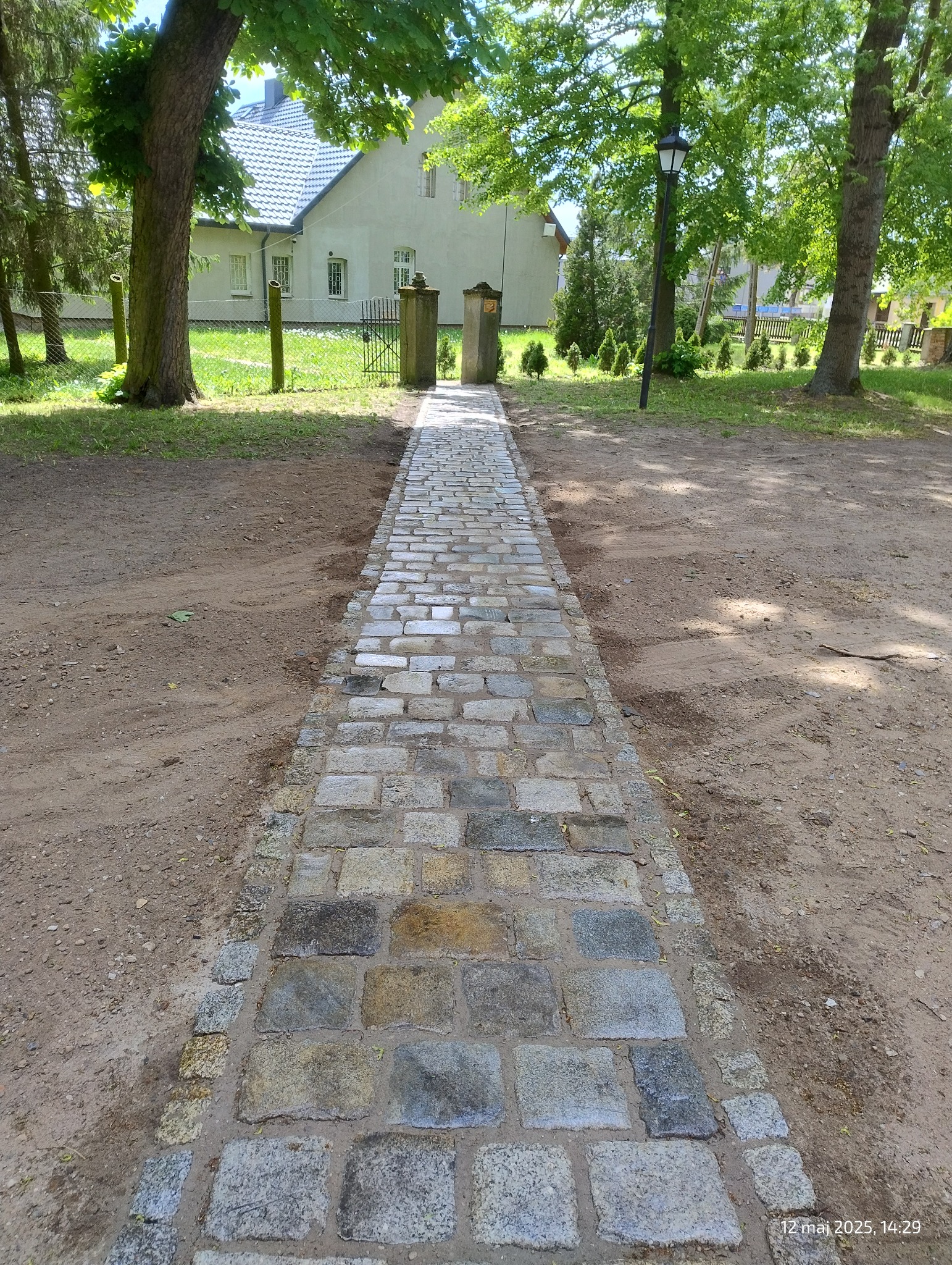 Ułożona z kamiennej kostki ścieżka prowadzi przez trawnik do bramki w ogrodzeniu przed domem. Widoczne drzewa i latarnia. Kostka brukowa w odcieniach szarości i beżu.