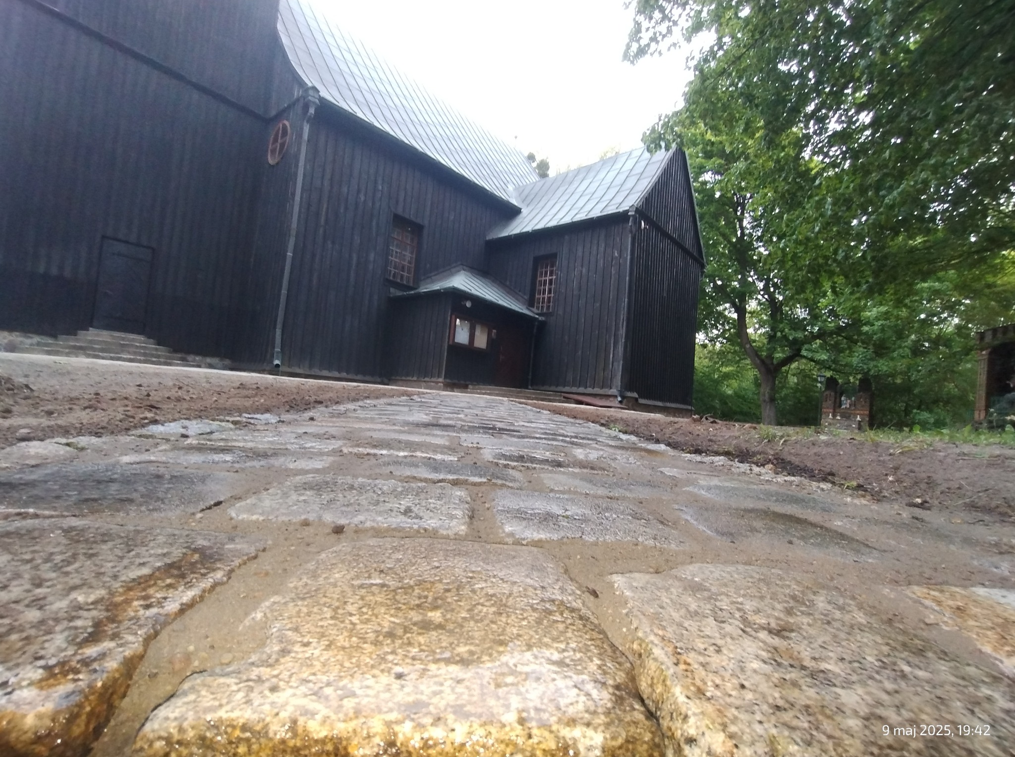 Ułożona, kamienna droga prowadzi do drewnianego kościoła o ciemnej elewacji. Widok z niskiej perspektywy podkreśla fakturę kamieni i prowadzi wzrok do budynku.