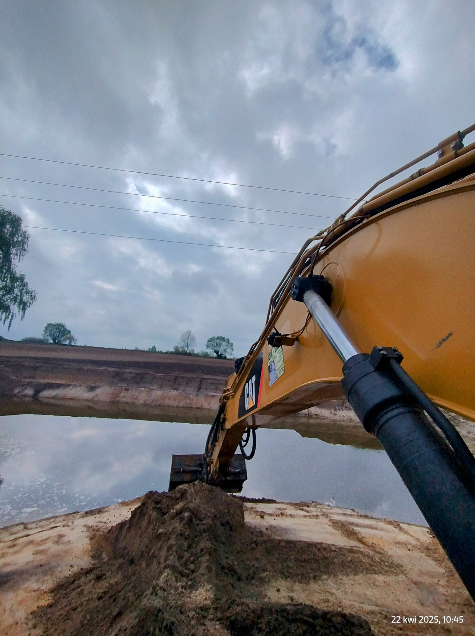 Żółta koparka CAT przy wykopanym stawie, pochmurne niebo odbija się w wodzie. Widok z dołu na ramię maszyny i wykopany brzeg.