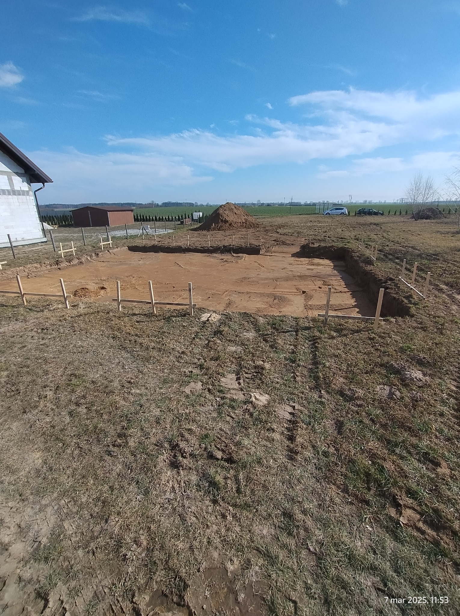 Wykonany wykop pod fundamenty z drewnianym szalunkiem, widok z góry na brązową ziemię i błękitne niebo. W tle budynek w stanie surowym i sterta ziemi.