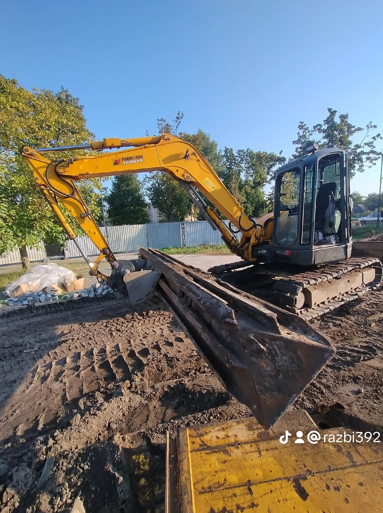 Żółta koparka AMMANN YANMAR na placu budowy, z łyżką skierowaną w stronę ziemi. W tle widoczne drzewa i metalowe ogrodzenie. Na ziemi leżą kostki brukowe.