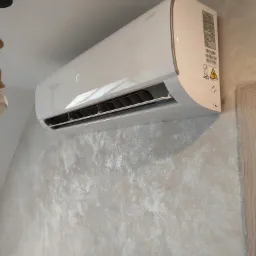 Biały klimatyzator ścienny marki Ukura System zamontowany na teksturowanej, jasnej ścianie w pomieszczeniu.