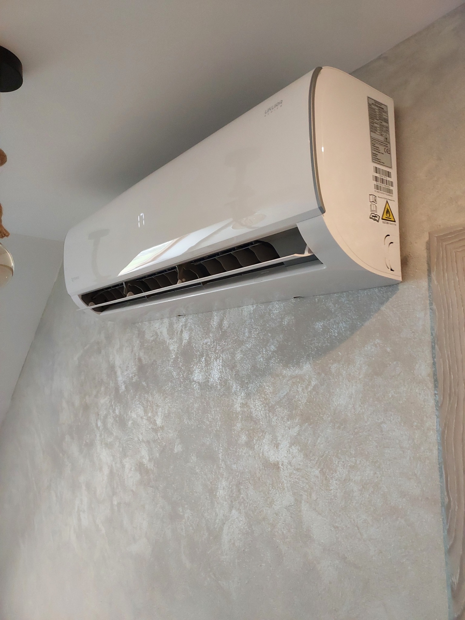 Biały klimatyzator ścienny marki Ukura System zamontowany na teksturowanej, jasnej ścianie w pomieszczeniu.