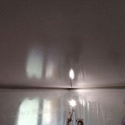 DAS the art of real estate sp. zo.o. - Świeżo pomalowany sufit w pomieszczeniu zabezpieczonym folią malarską, widoczne lampy oświetlające powierzchnię i fragment sylwetki malarza w masce ochronnej.