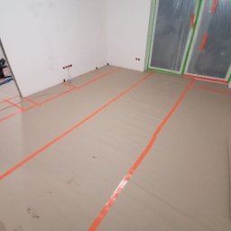 DAS the art of real estate sp. zo.o. - Przygotowanie pomieszczenia do prac wykończeniowych: podłoga zabezpieczona kartonem i taśmą malarską, okna ofoliowane, widoczny odkurzacz przemysłowy w rogu pomieszczenia.