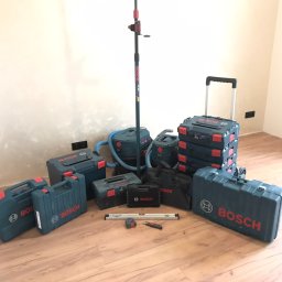 DAS the art of real estate sp. zo.o. - Zestaw narzędzi i akcesoriów marki Bosch, w tym skrzynki narzędziowe, odkurzacze przemysłowe i niwelator laserowy na statywie, rozłożone na podłodze z paneli w pomieszczeniu z surowymi ścianami...