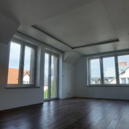 DAS the art of real estate sp. zo.o. - Puste, jasne poddasze z ciemną drewnianą podłogą i białym sufitem z dekoracyjną wnęką, widok na zewnątrz przez okna.