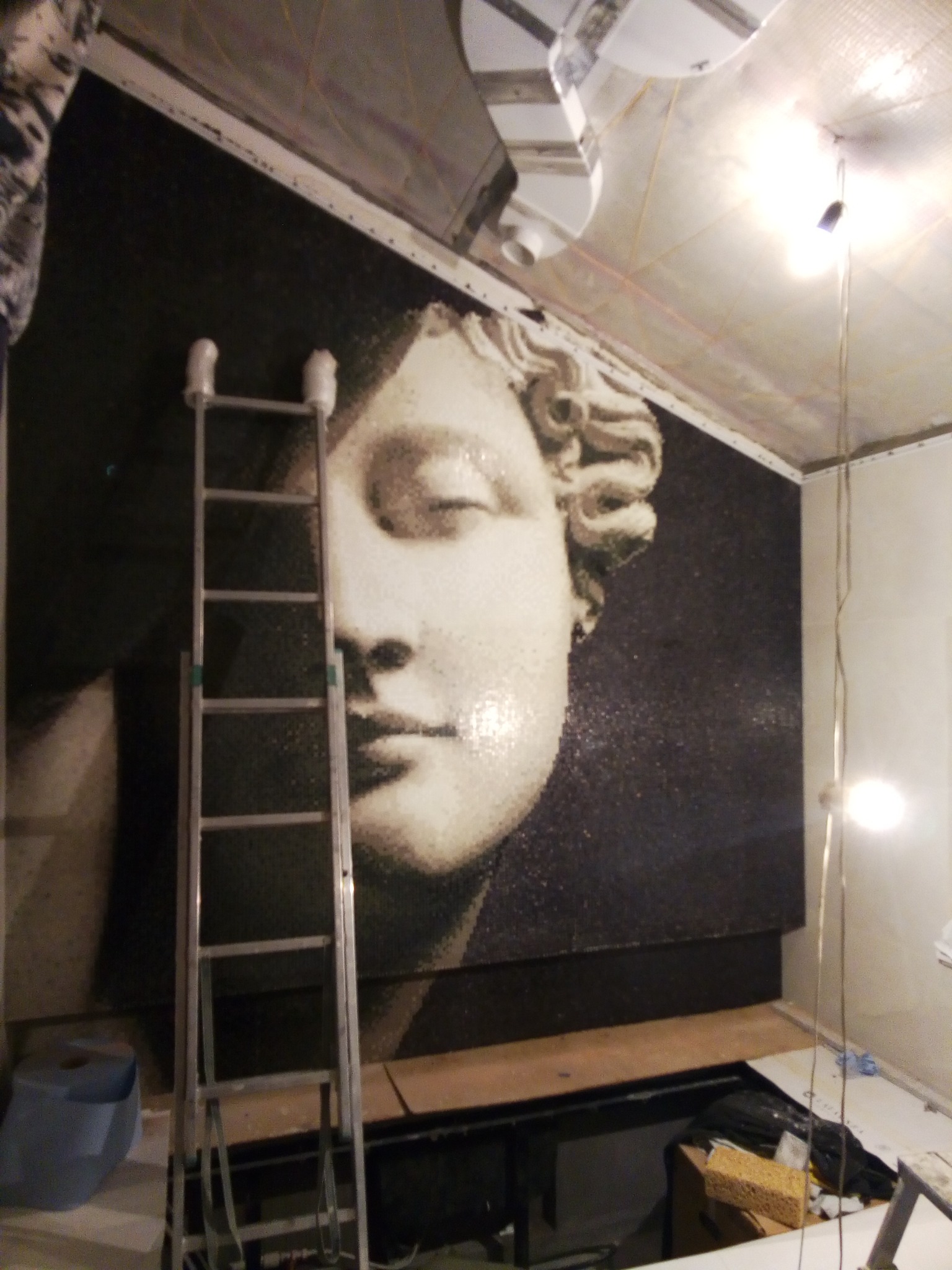 Mozaika z wizerunkiem twarzy kobiety w stylu antycznym na ścianie, widoczna drabina aluminiowa i elementy instalacji sufitowej.